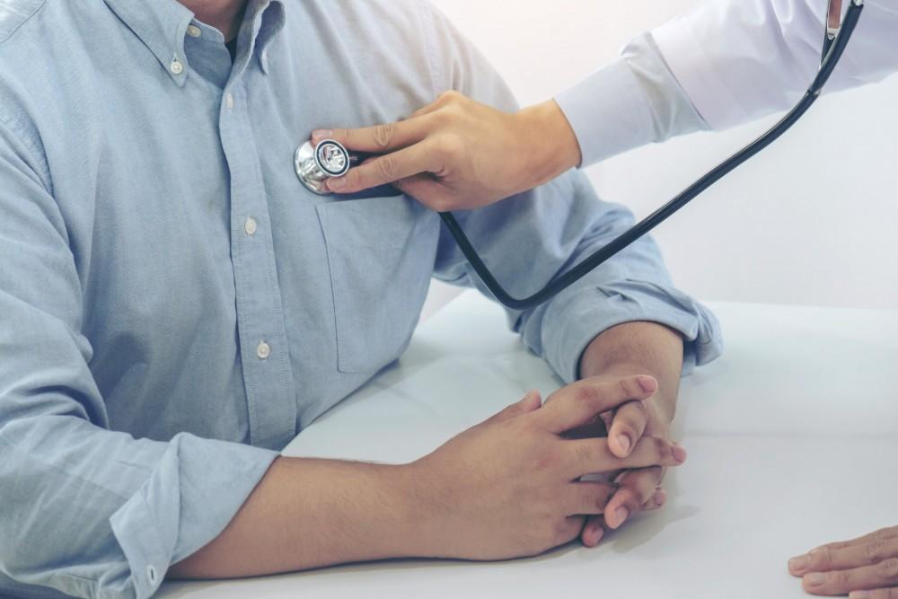 A doctor uses a stethoscope to listen to the chest of a patient wearing a light blue shirt, while the patient sits with their hands folded on a white table.