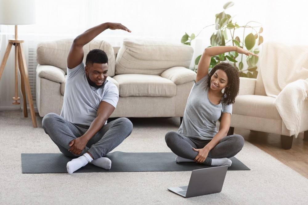 Two people sit on yoga mats in a living room, stretching with one arm overhead and smiling while following an exercise video on a laptop. There is a couch and plants in the background.