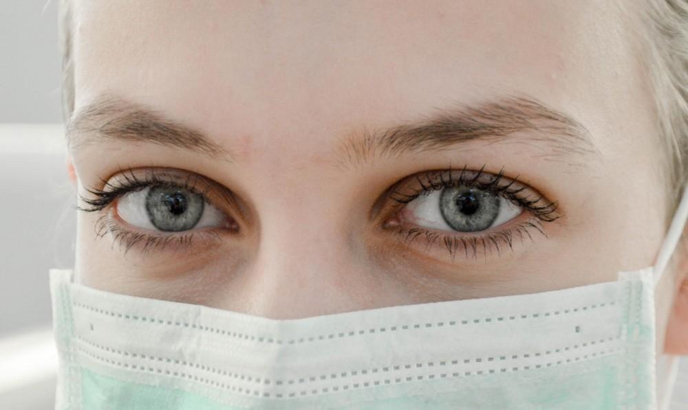 Close-up of a persons blue eyes and eyebrows, with the rest of their face covered by a light green surgical mask. The background is out of focus.