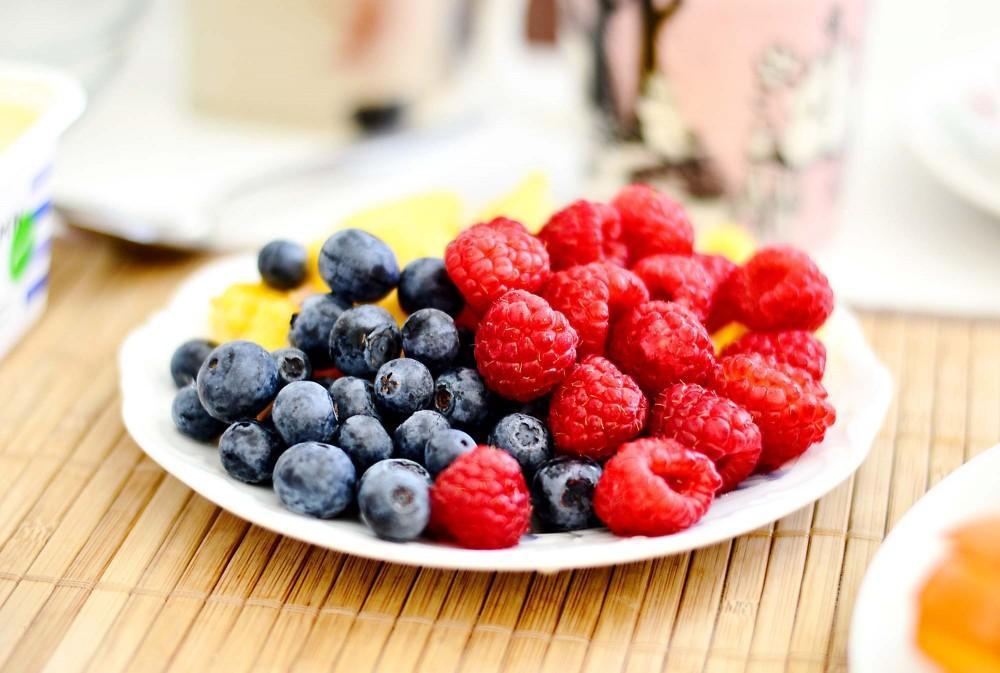 A white plate filled with fresh blueberries and raspberries sits on a bamboo mat. Some small pieces of yellow fruit are visible in the background.