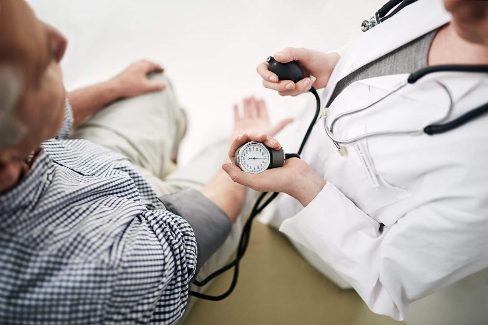 A healthcare professional in a white coat measures an older mans blood pressure using a sphygmomanometer and stethoscope. The man is seated, and the scene is viewed from above.