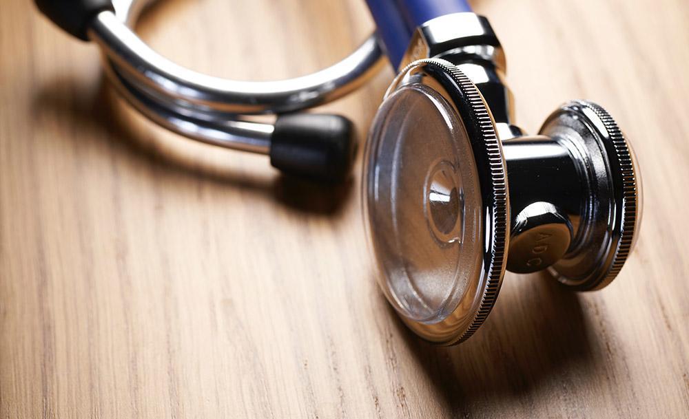A close-up of a stethoscope lying on a wooden surface, showing the shiny metal chest piece and part of the blue tubing.