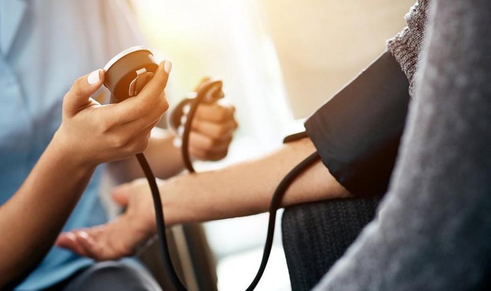 A healthcare professional uses a blood pressure monitor to check a persons blood pressure. Sunlight streams in the background, creating a warm atmosphere. Only the hands and arms of both individuals are visible.