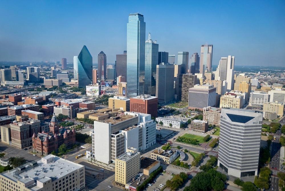 Aerial view of downtown Dallas, Texas, showing a mix of modern skyscrapers, including tall glass buildings, and historic structures under a clear blue sky.
