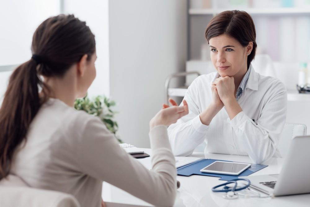 A doctor attentively listens to a patient during a consultation in a bright office. Both women are seated at a desk with paperwork, a tablet, and a stethoscope visible.