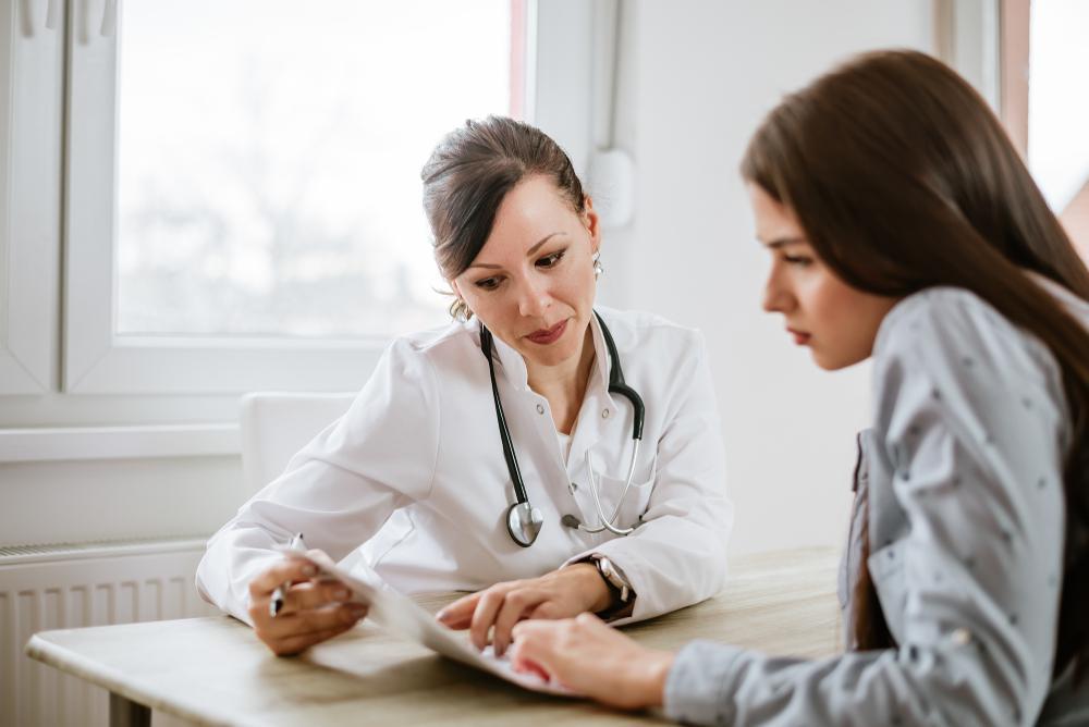 A female doctor with a stethoscope discusses paperwork with a female patient at a desk in a bright, modern office. The doctor is explaining or reviewing a document with the patient.