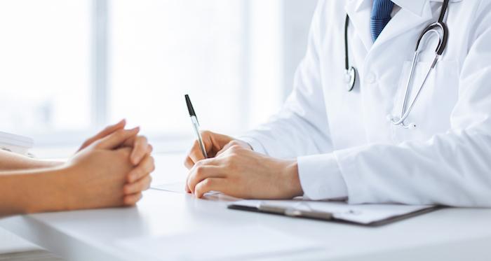 A doctor in a white coat with a stethoscope consults a patient, holding a pen and clipboard, while the patient sits across the desk with hands clasped.