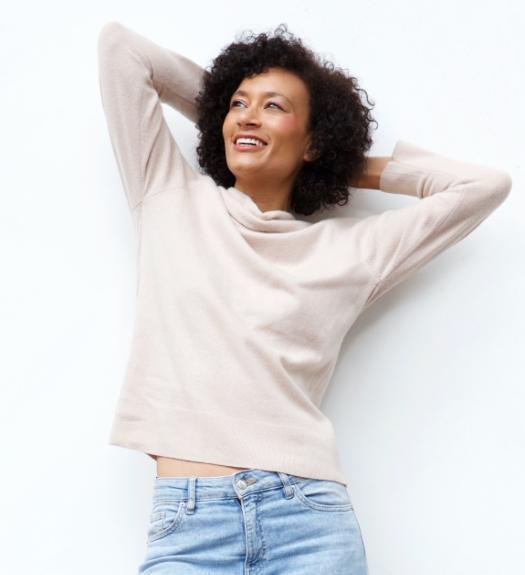 A smiling woman with curly hair wears a light beige sweater and light blue jeans, standing against a white background with her arms raised and hands behind her head.