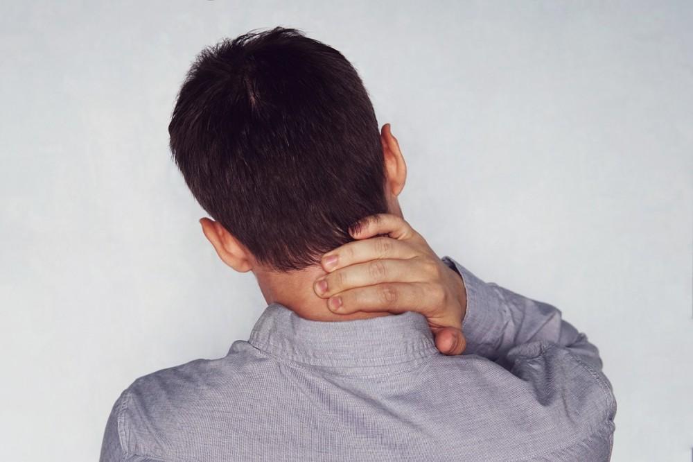 A person seen from behind, wearing a light gray shirt, holds the back of their neck with one hand, suggesting discomfort or pain.
