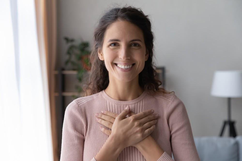 A woman with long, wavy brown hair smiles warmly while placing both hands over her chest. She is wearing a light pink ribbed top and standing indoors, with blurred plants and furniture in the background.