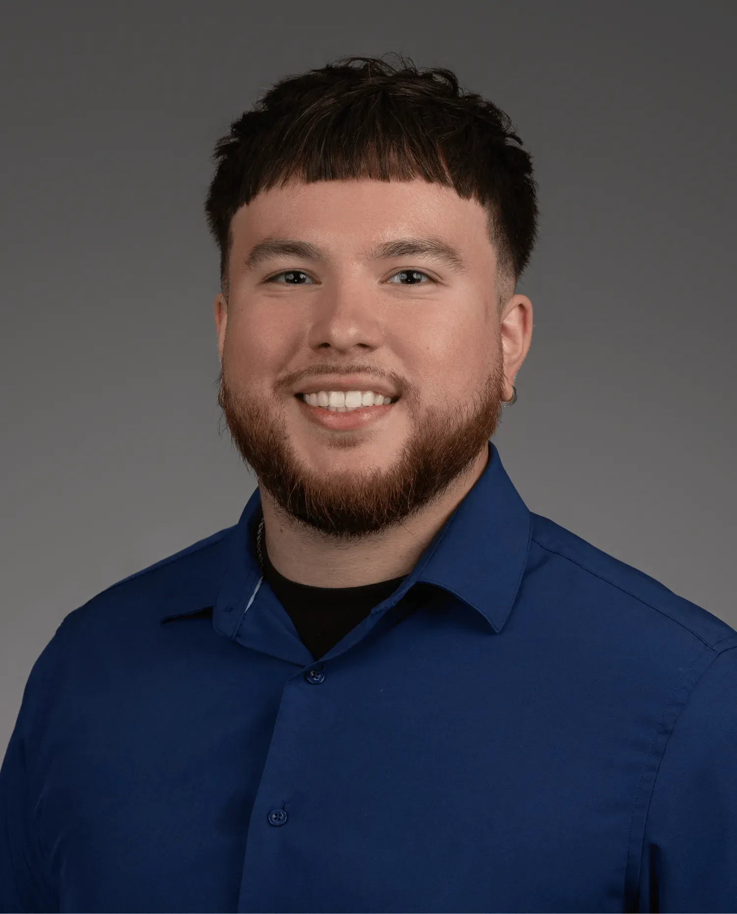 A young man with short brown hair, a beard, and mustache is smiling. He is wearing a blue collared shirt over a black t-shirt. The background is plain gray.