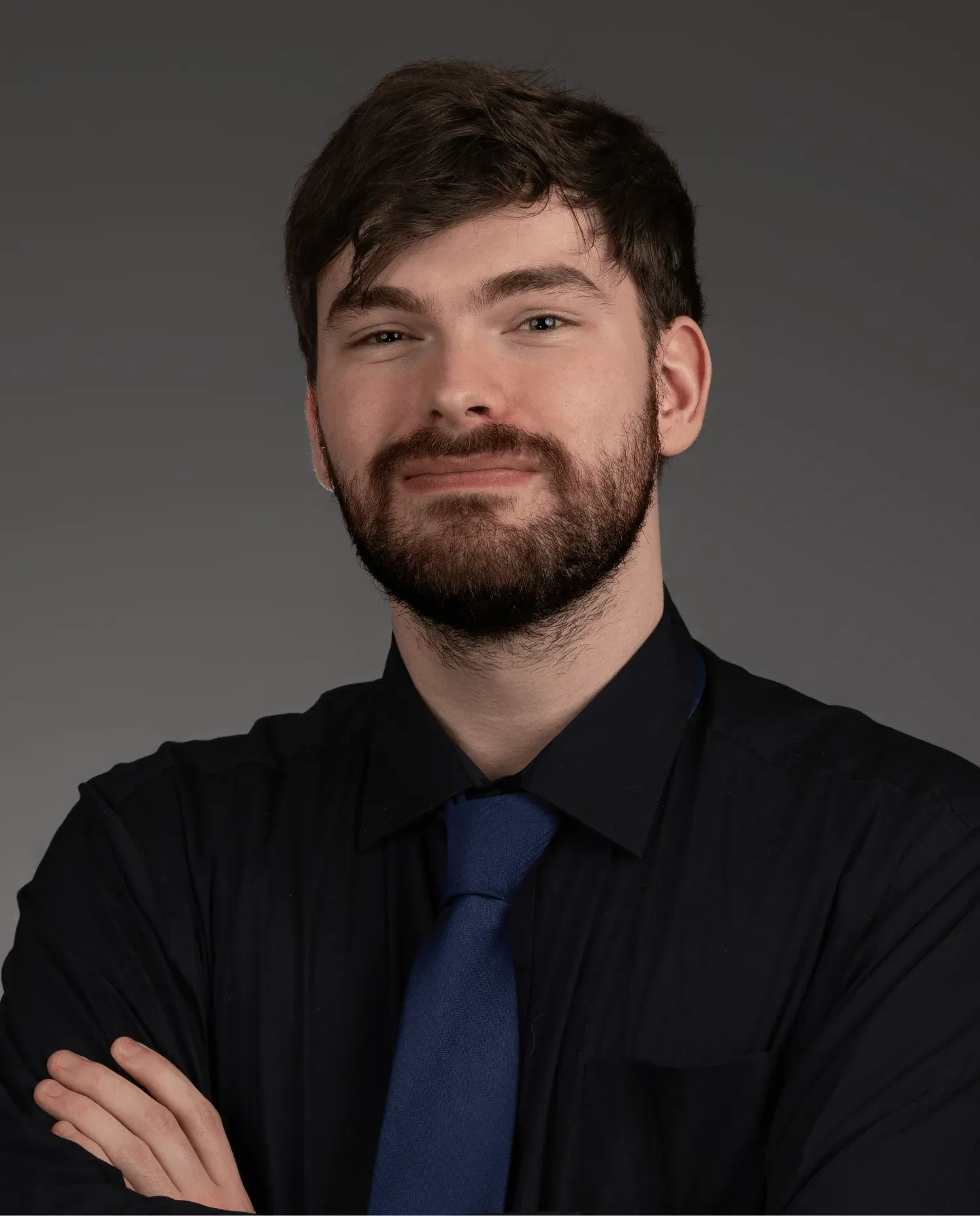 A young man with a beard and short brown hair, wearing a dark shirt and blue tie, stands with arms crossed and smiles slightly against a plain gray background.