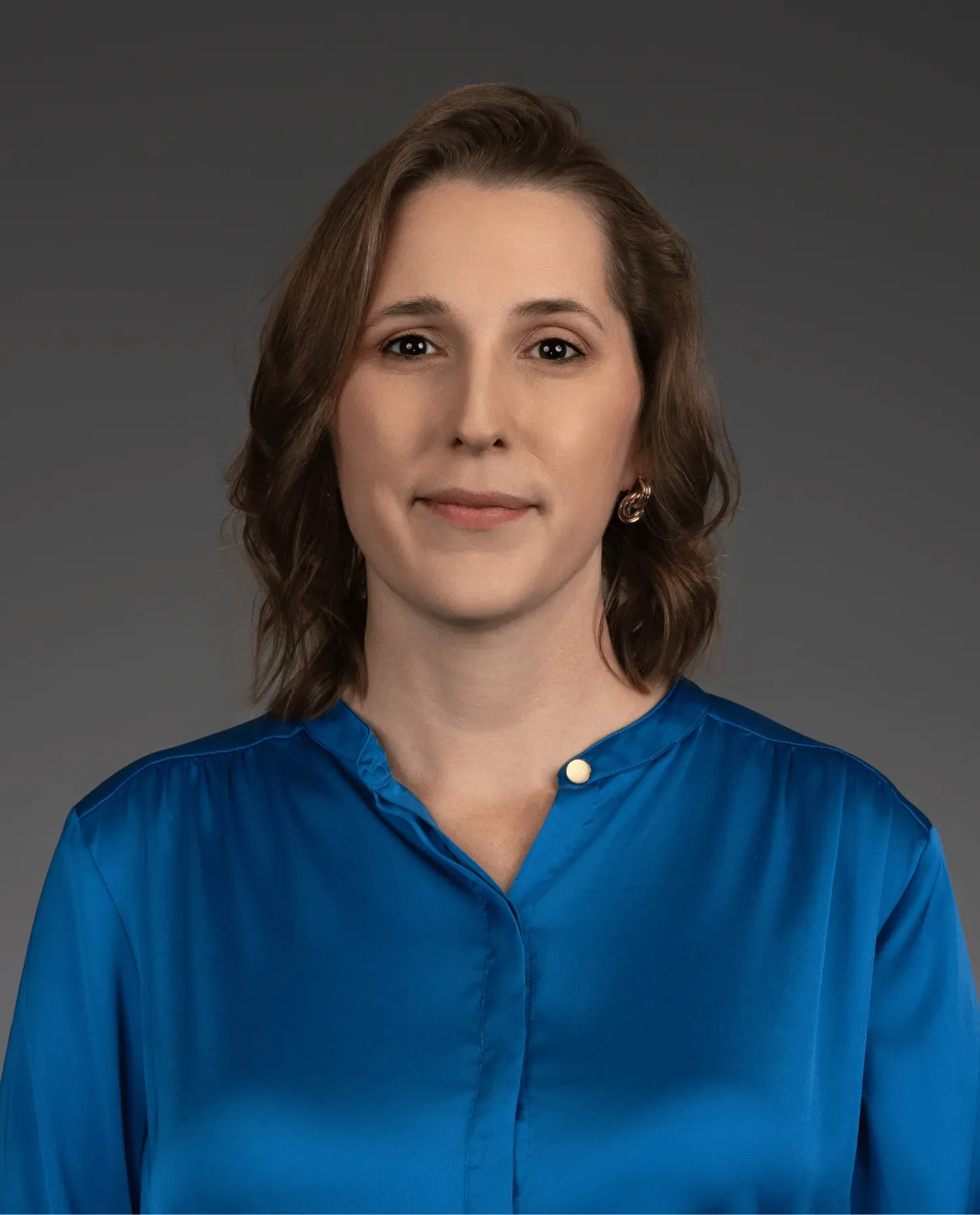 A woman with shoulder-length brown hair wearing a royal blue blouse stands against a plain gray background, looking directly at the camera with a neutral expression.