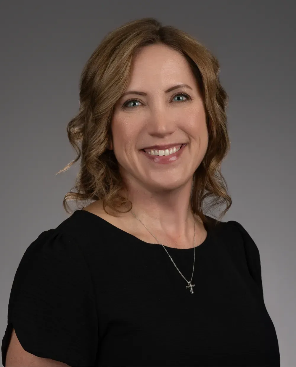 A woman with wavy, shoulder-length light brown hair smiles at the camera. She is wearing a black top, a silver necklace with a cross pendant, and stands against a plain gray background.