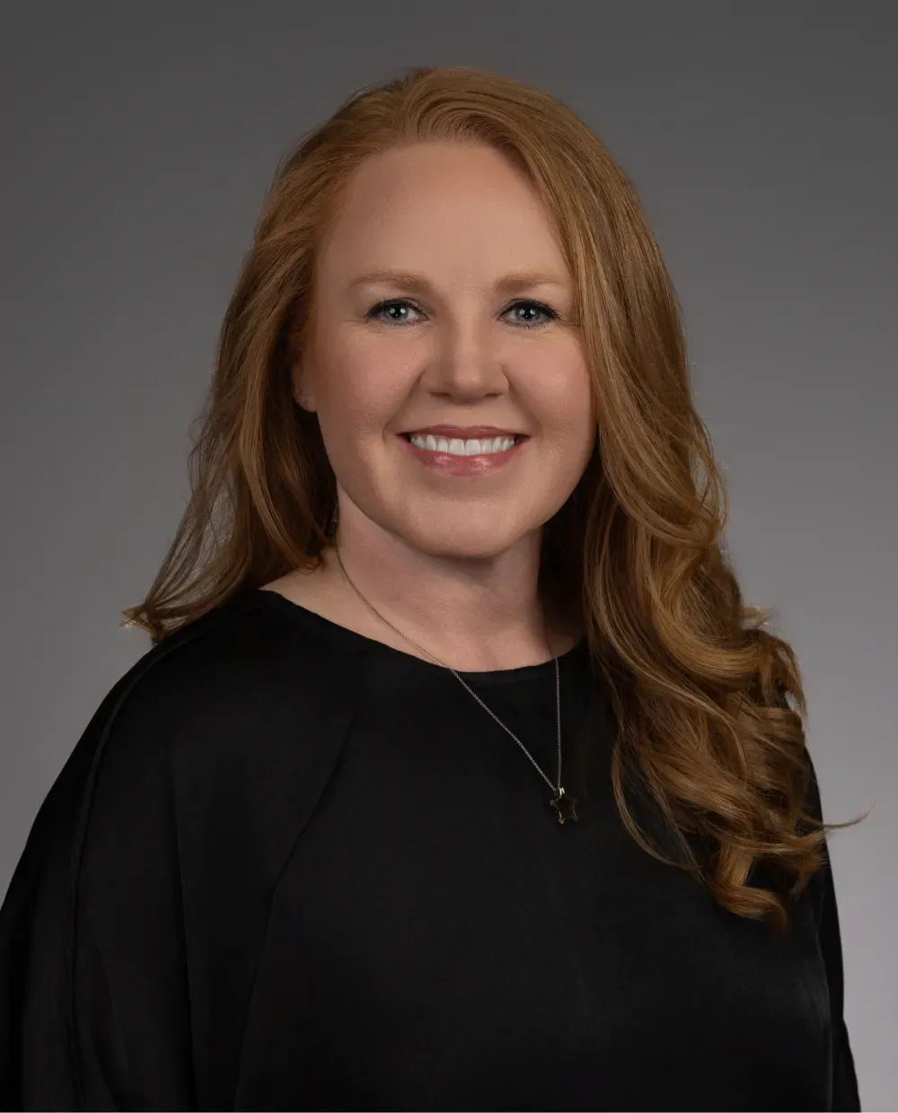 A woman with long, wavy red hair is smiling at the camera. She is wearing a black top and a necklace with a small pendant, posed against a plain gray background.