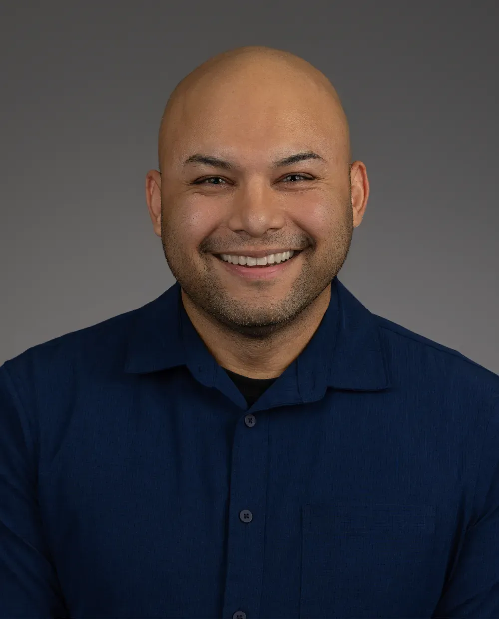 Smiling man with a shaved head wearing a navy blue button-up shirt, posed against a plain gray background.