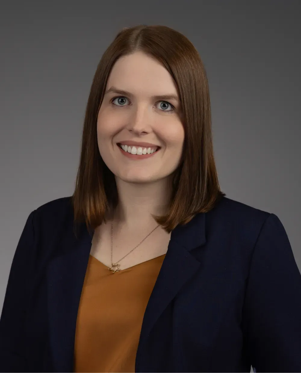 A woman with straight, shoulder-length brown hair, wearing a navy blazer over a mustard-colored top, smiles at the camera against a plain gray background.