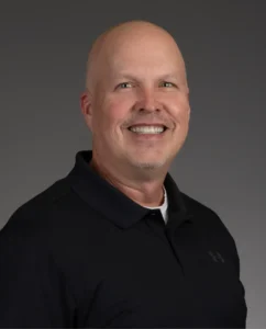 Smiling bald man wearing a black polo shirt poses in front of a plain gray background.