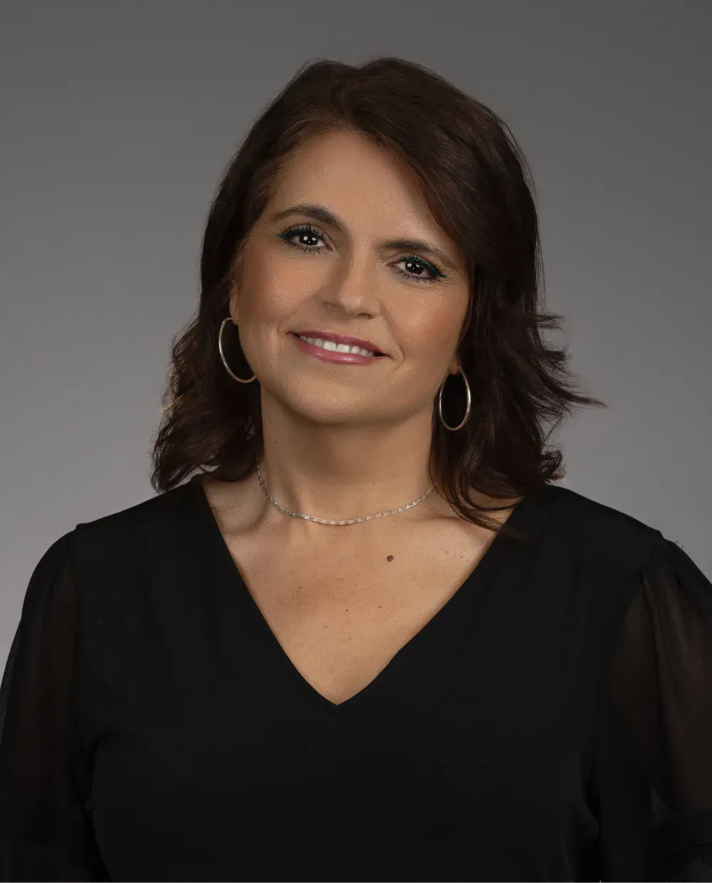 A woman with medium-length brown hair, wearing hoop earrings, a delicate necklace, and a black V-neck top, smiles softly against a plain gray background.