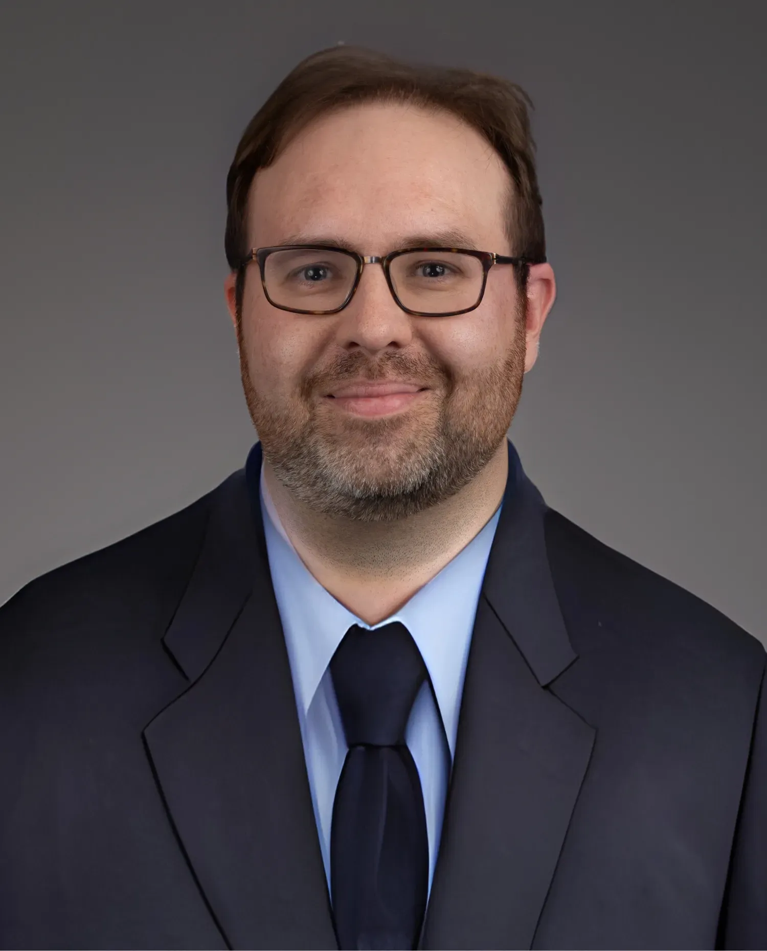 A man with short brown hair, a beard, and glasses is wearing a dark suit, light blue shirt, and dark tie, smiling slightly in front of a plain gray background.