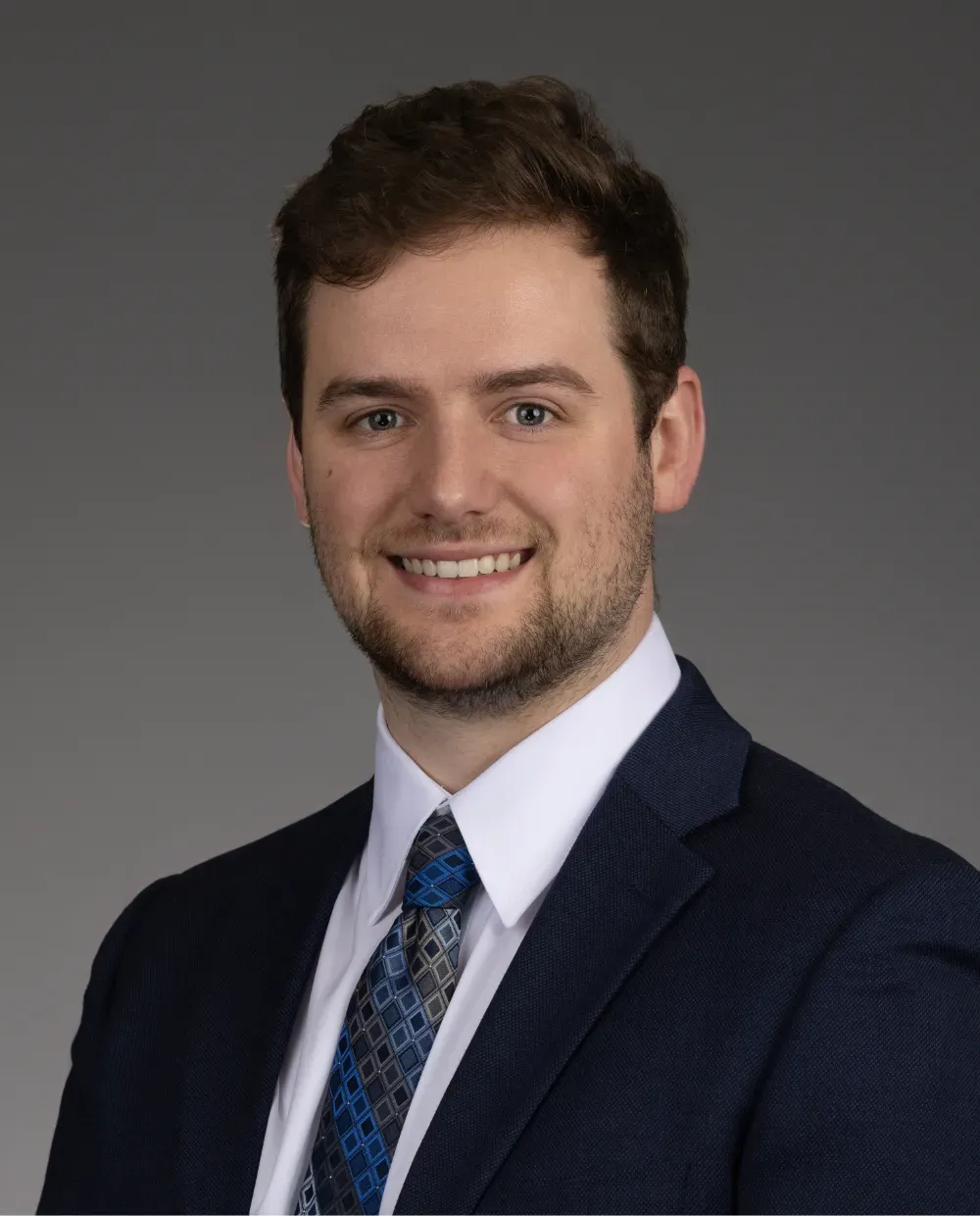 A young man with short brown hair and a trimmed beard, wearing a navy suit, white dress shirt, and patterned blue tie, smiles in a professional studio portrait against a plain gray background.