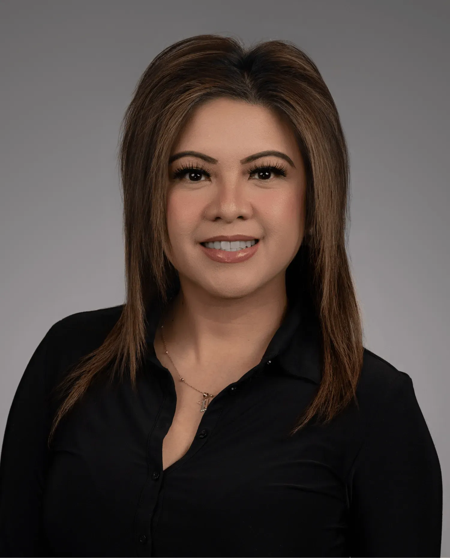 A woman with straight, shoulder-length brown hair, wearing a black collared shirt and a necklace, smiles at the camera against a plain gray background.