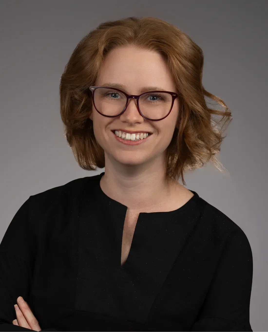 A woman with shoulder-length wavy red hair, wearing glasses and a black top, smiles at the camera against a plain gray background.