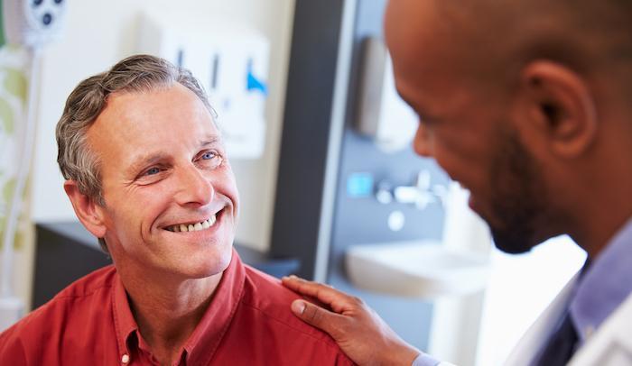 A doctor places a reassuring hand on a smiling middle-aged man’s shoulder during a conversation in a medical office.