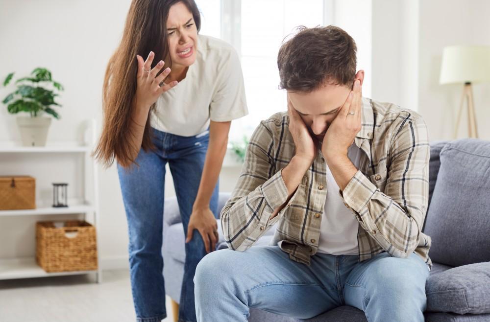 A woman stands and gestures angrily at a man sitting on a couch with his head in his hands, looking stressed or upset in a bright living room.