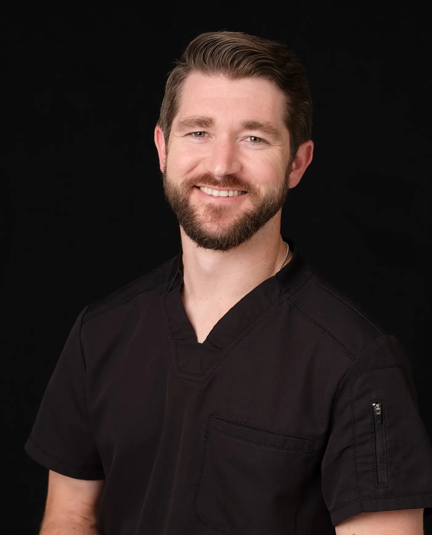 A man with short brown hair and a beard, wearing black medical scrubs, smiles at the camera against a plain black background.