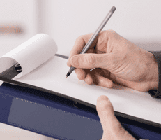 A person holds a clipboard and writes on a sheet of paper with a pen, suggesting note-taking or form filling.