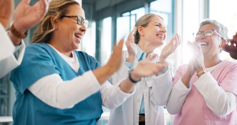A group of smiling healthcare professionals in scrubs and lab coats stand together indoors, clapping and celebrating, with sunlight streaming through large windows behind them.