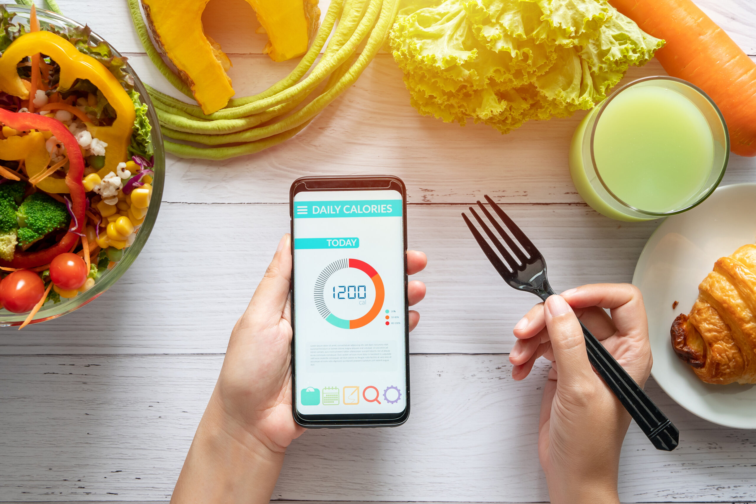 A person holds a smartphone displaying a daily calories app and a fork, surrounded by a salad, croissant, green juice, fresh vegetables, and lettuce on a white wooden table.