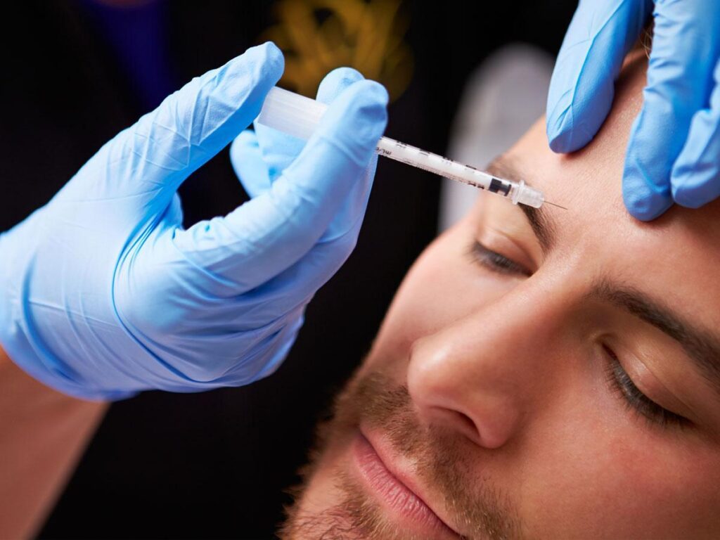 A close-up of a man receiving a cosmetic injection in his forehead. A person wearing blue gloves holds his head and administers the injection with a syringe.