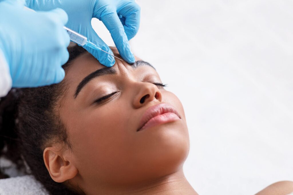 A person with closed eyes receives an injection between the eyebrows from a gloved medical professional, possibly for a cosmetic procedure like Botox, while lying down.