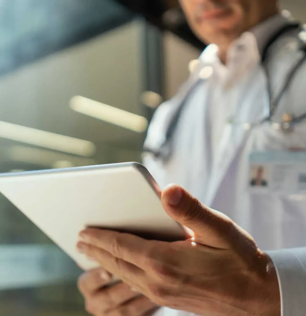 A doctor wearing a white coat and stethoscope holds and uses a tablet device, standing indoors with blurred background.