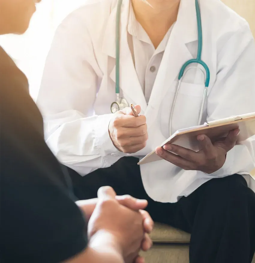 A doctor with a stethoscope around their neck holds a clipboard and discusses with a patient, who sits with hands clasped, during a consultation.