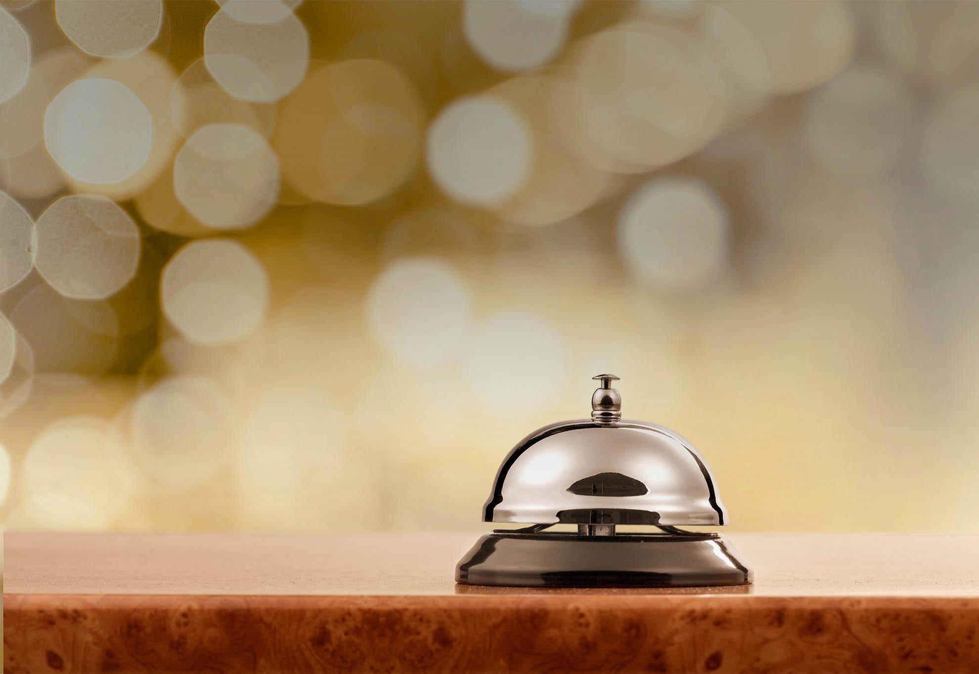 A shiny silver service bell sits on a wooden counter with a blurred golden bokeh background, creating a warm and inviting atmosphere.