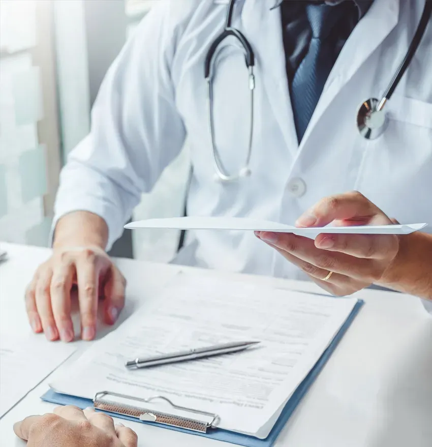 A doctor in a white coat with a stethoscope hands a document to someone, with a clipboard, pen, and medical forms on the desk in front of them.
