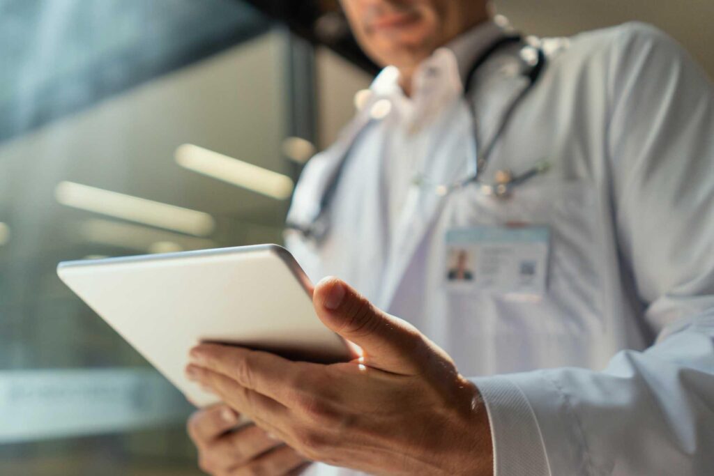 A doctor wearing a white lab coat and stethoscope uses a tablet device. The doctors face is out of focus, and sunlight streams in from a window in the background.