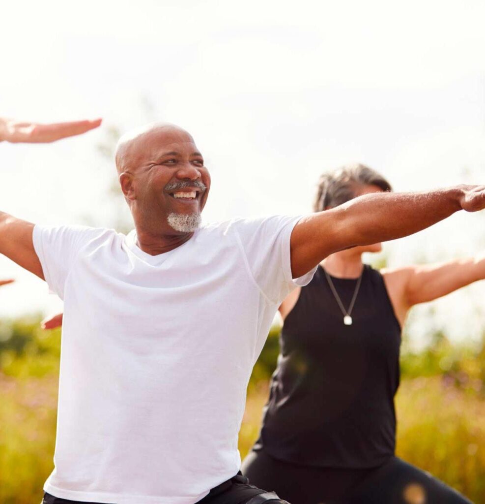 Two people practicing yoga outdoors. A smiling man in a white shirt is in the foreground with arms outstretched, performing a yoga pose. Another person in black is behind him, also participating. The background is blurred greenery.