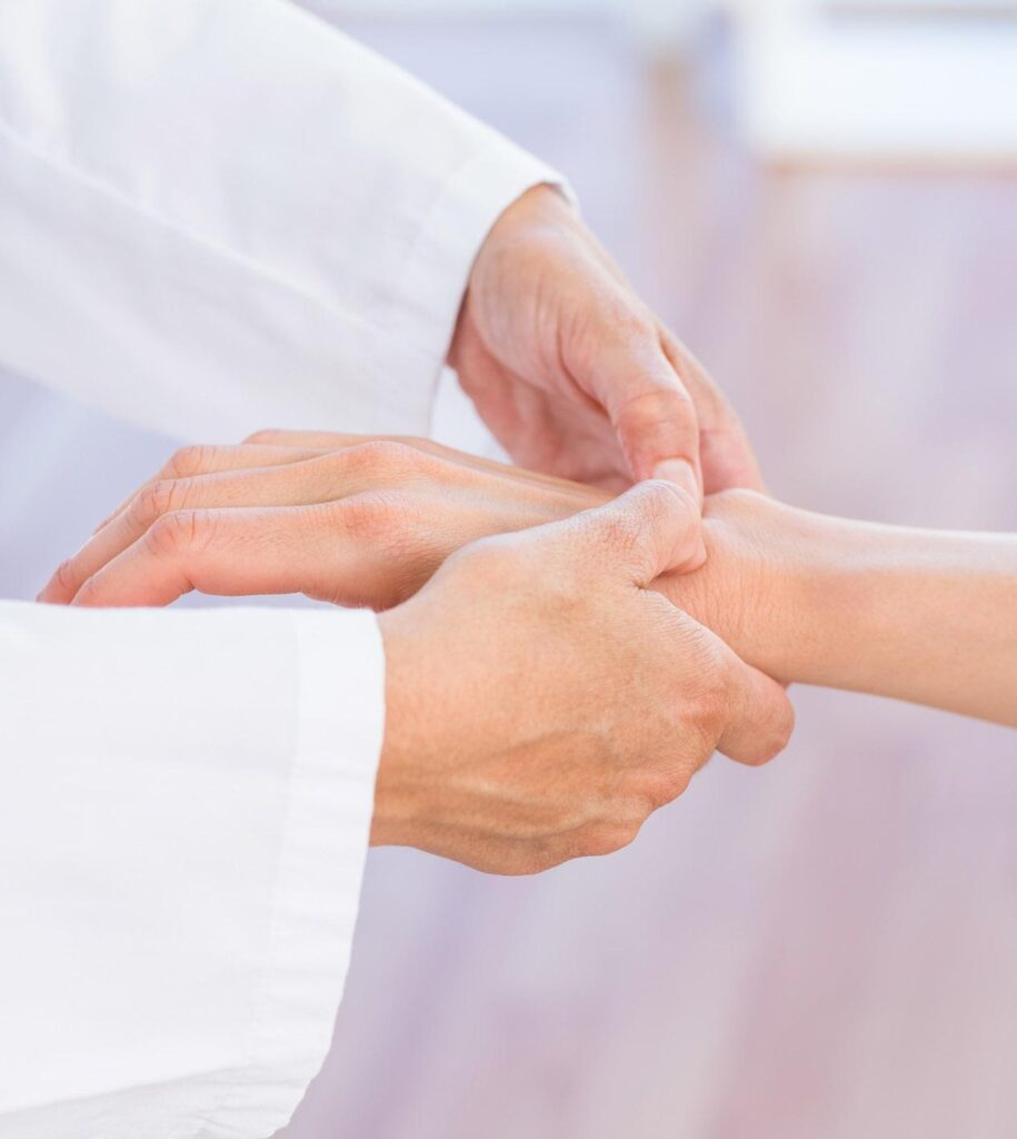 A healthcare professional gently holds and examines a persons wrist, possibly checking their pulse, with both individuals hands visible against a blurred background.