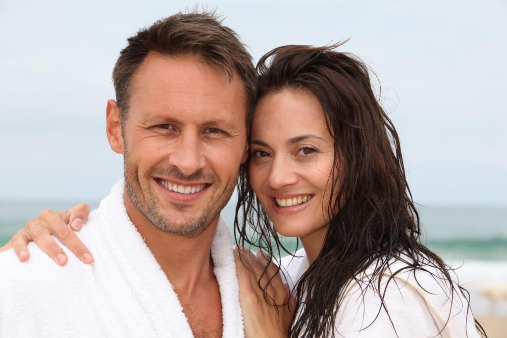 A smiling man and woman wearing white bathrobes stand close together on a beach, with the ocean and sky in the background. The woman has her arm around the man, and both have wet hair.