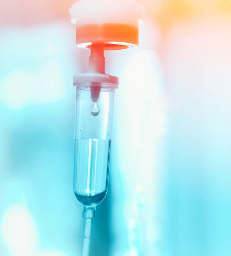 A close-up of a medical IV drip chamber with a droplet of fluid inside, set against a blurred blue and orange background. The image has a soft, glowing light effect.