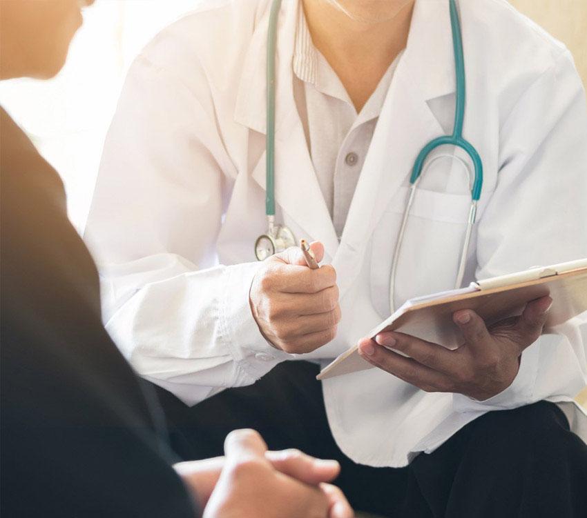 A doctor wearing a white coat and stethoscope holds a clipboard and pen, speaking with a seated patient whose face is not visible.