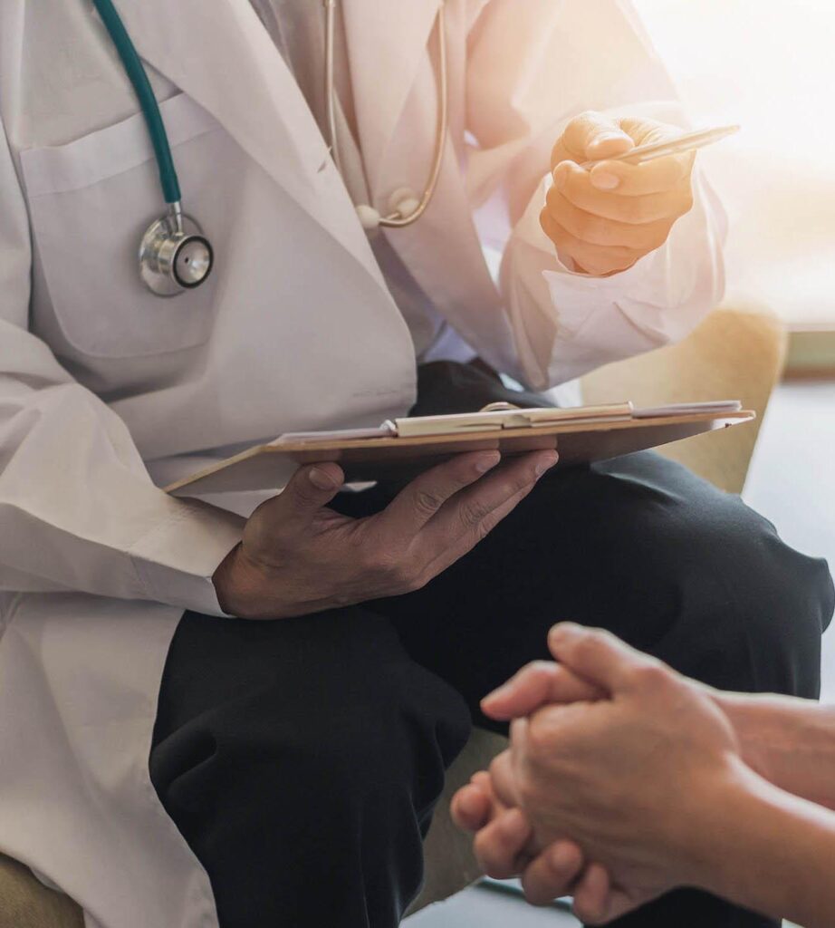 A doctor in a white coat with a stethoscope holds a clipboard and gestures while talking to a seated patient whose hands are clasped together.