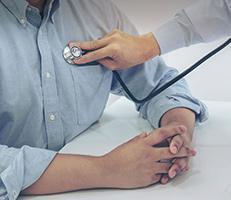 A healthcare professional uses a stethoscope to listen to a patients chest while the patient sits with hands folded on a white table.