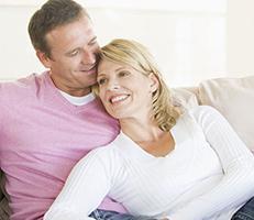 A smiling couple sits closely together on a couch. The woman, wearing a white top, leans against the man, who is wearing a pink shirt. They appear happy and relaxed in a bright, cozy room.