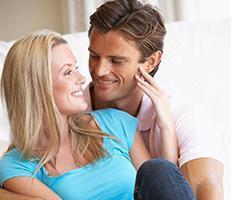 A smiling couple sits close together on a couch, looking into each other’s eyes. The woman has blonde hair and wears a blue shirt, while the man has brown hair and wears a light pink shirt.