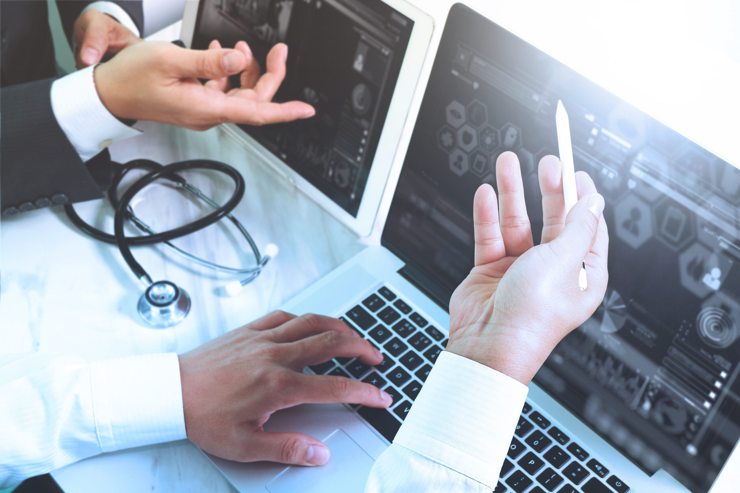 Two people discuss medical data on a laptop and tablet. One types on the laptop, while the other gestures with a pen. A stethoscope sits on the desk, suggesting a healthcare or medical setting.